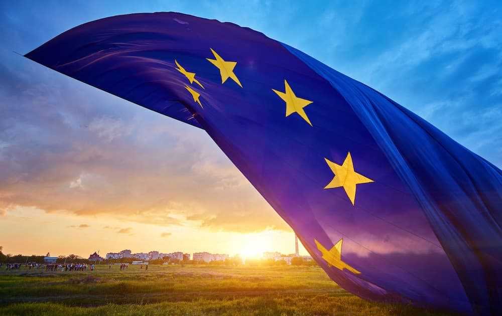 European Union flag with stars waving against a sunset sky with clouds. - Olive Oil Times