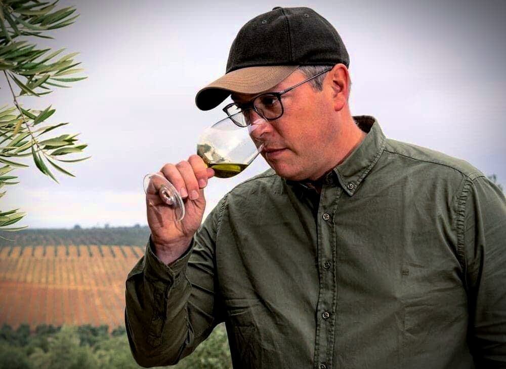 Moški v zeleni srajci in kapici okuša oljčno olje iz kozarca v vinogradu. - Olive Oil Times