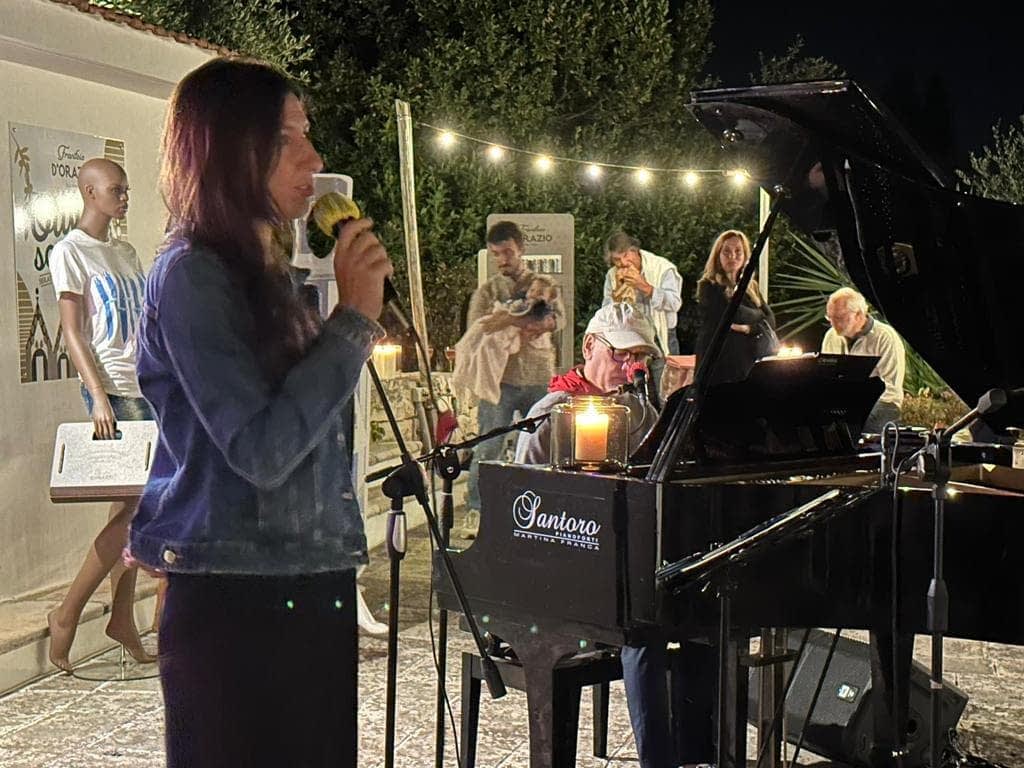 A woman singing into a microphone while seated at a piano during a performance at night. - Olive Oil Times