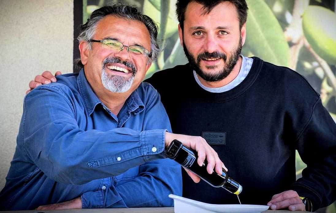 Zwei Männer lächeln, während einer von ihnen Olivenöl aus einer Flasche in eine Schüssel gießt. - Olive Oil Times