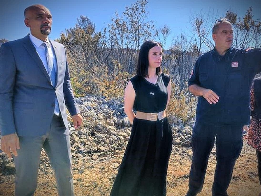 Three individuals standing outdoors, two in formal attire and one in work uniform, engaged in conversation. - Olive Oil Times