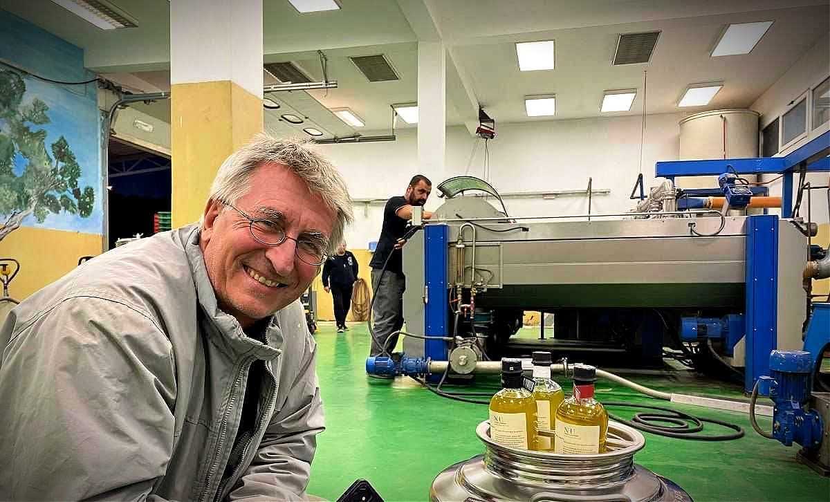 Un uomo sorridente in un impianto di lavorazione dell'olio d'oliva con macchinari e bottiglie di olio d'oliva in primo piano. - Olive Oil Times