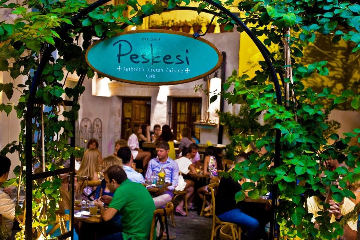 Eingangsschild des Restaurants Peskesi mit Grünflächen und Gästen, die drinnen speisen. – Olive Oil Times