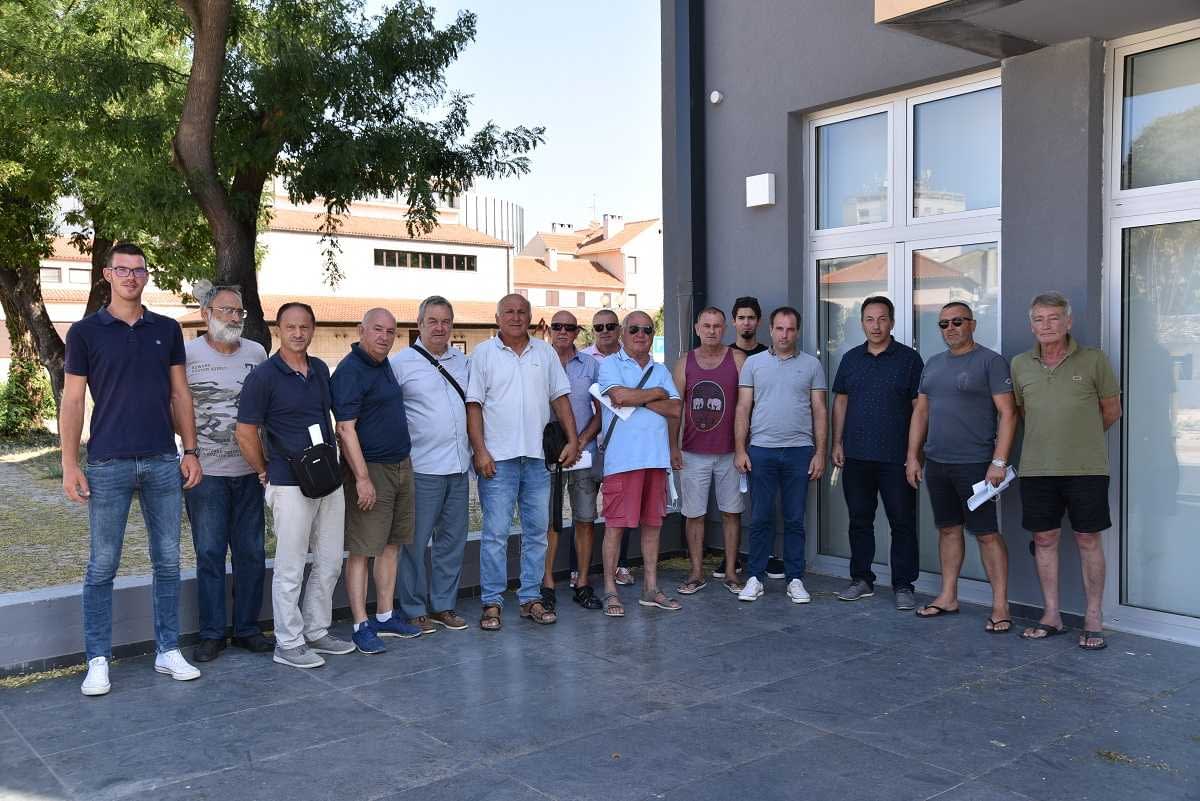 A group of individuals standing together in a line outside a building, some wearing sunglasses and casual clothing. - Olive Oil Times