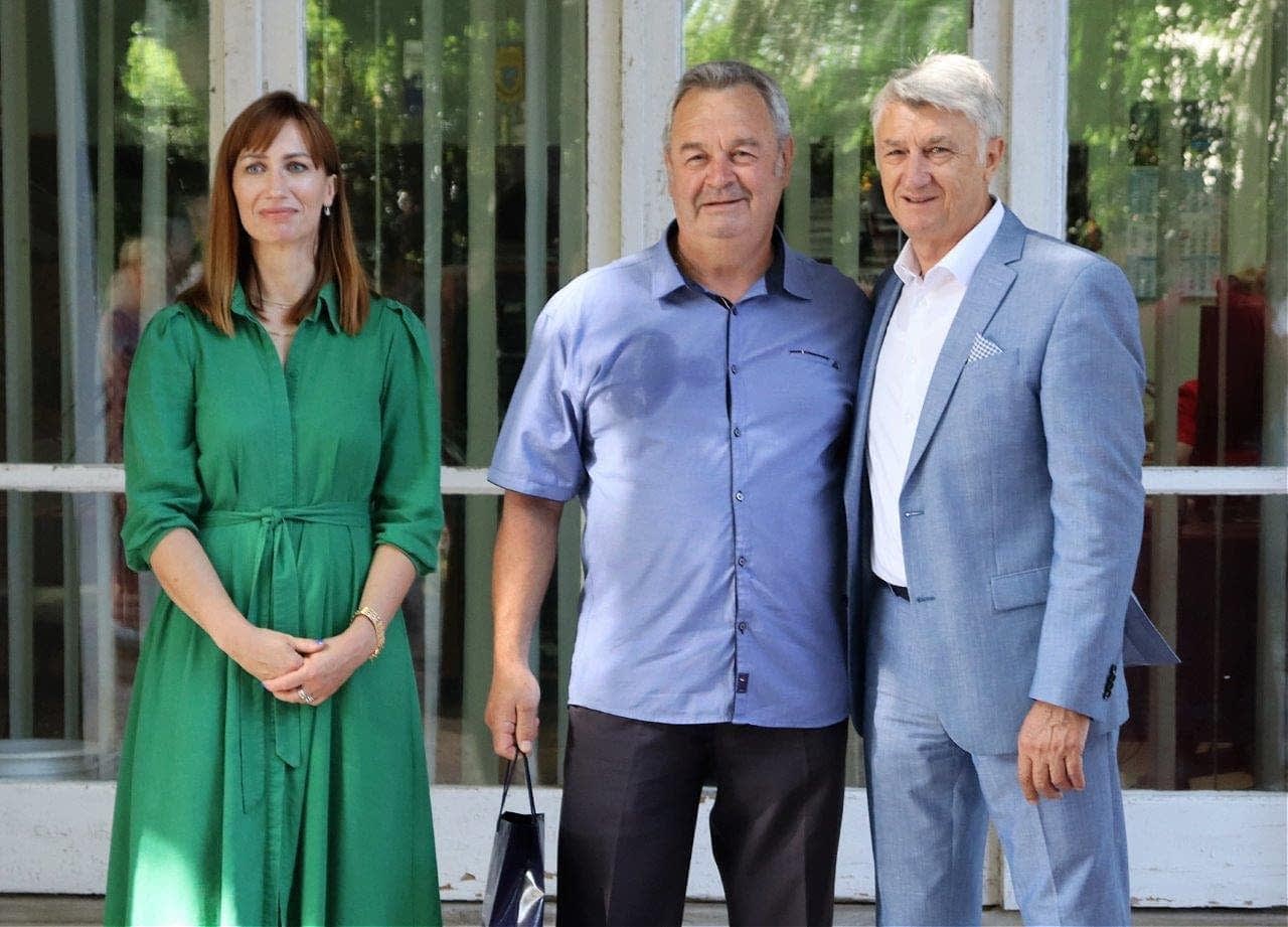 Three individuals standing together for a photo, with one woman in a green dress and two men in casual and formal attire. - Olive Oil Times