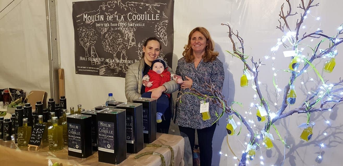 Two women and a baby at an olive oil stand with products displayed in a tent. - Olive Oil Times