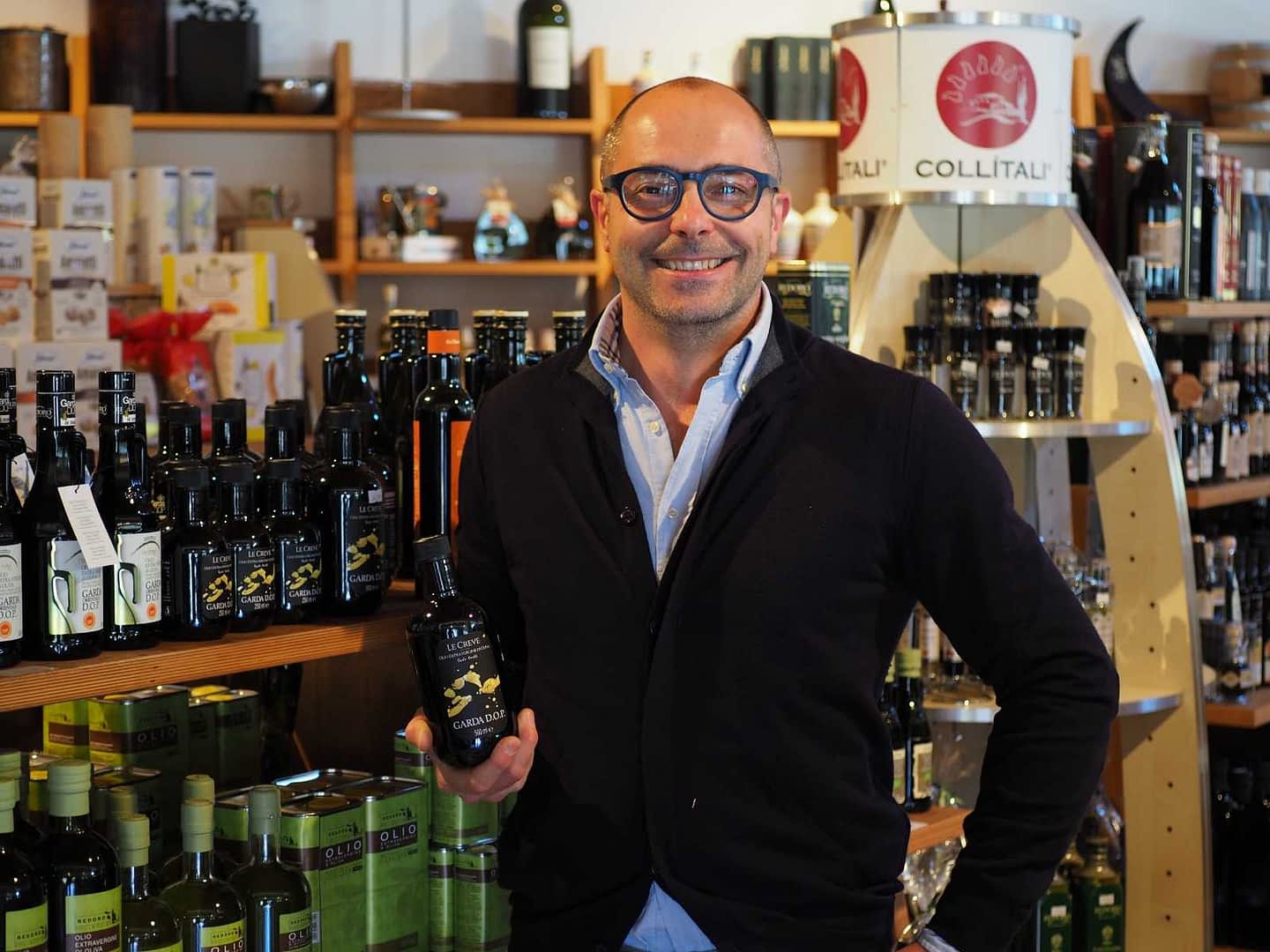 Un homme avec des lunettes tenant une bouteille d'huile d'olive dans un magasin rempli de divers produits pétroliers. - Olive Oil Times