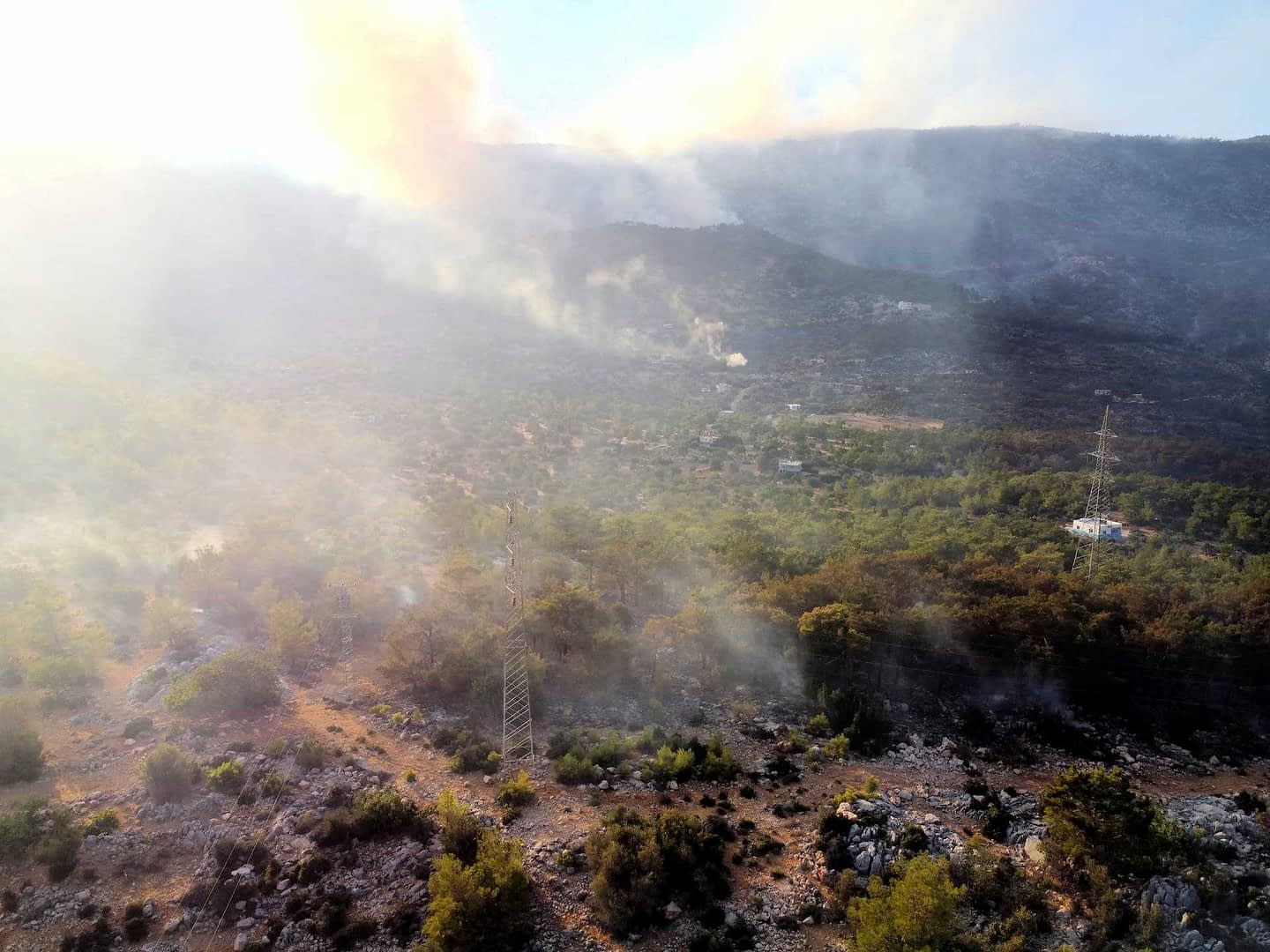 Vue aérienne d'un paysage avec de la fumée s'élevant du sol parmi les arbres et le terrain rocheux. - Olive Oil Times