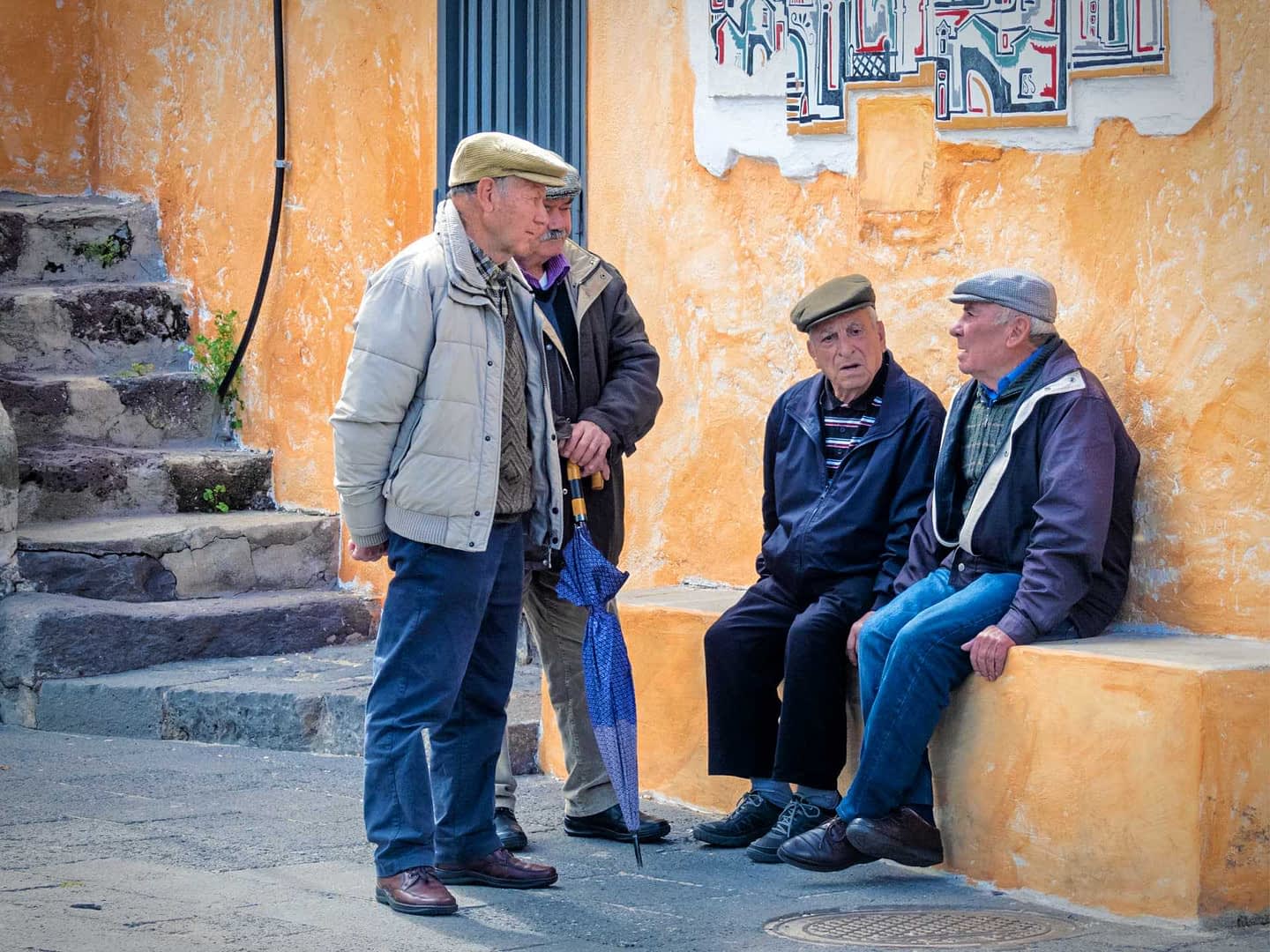 Four elderly men wearing caps engaged in conversation while seated and standing near a colorful wall. - Olive Oil Times
