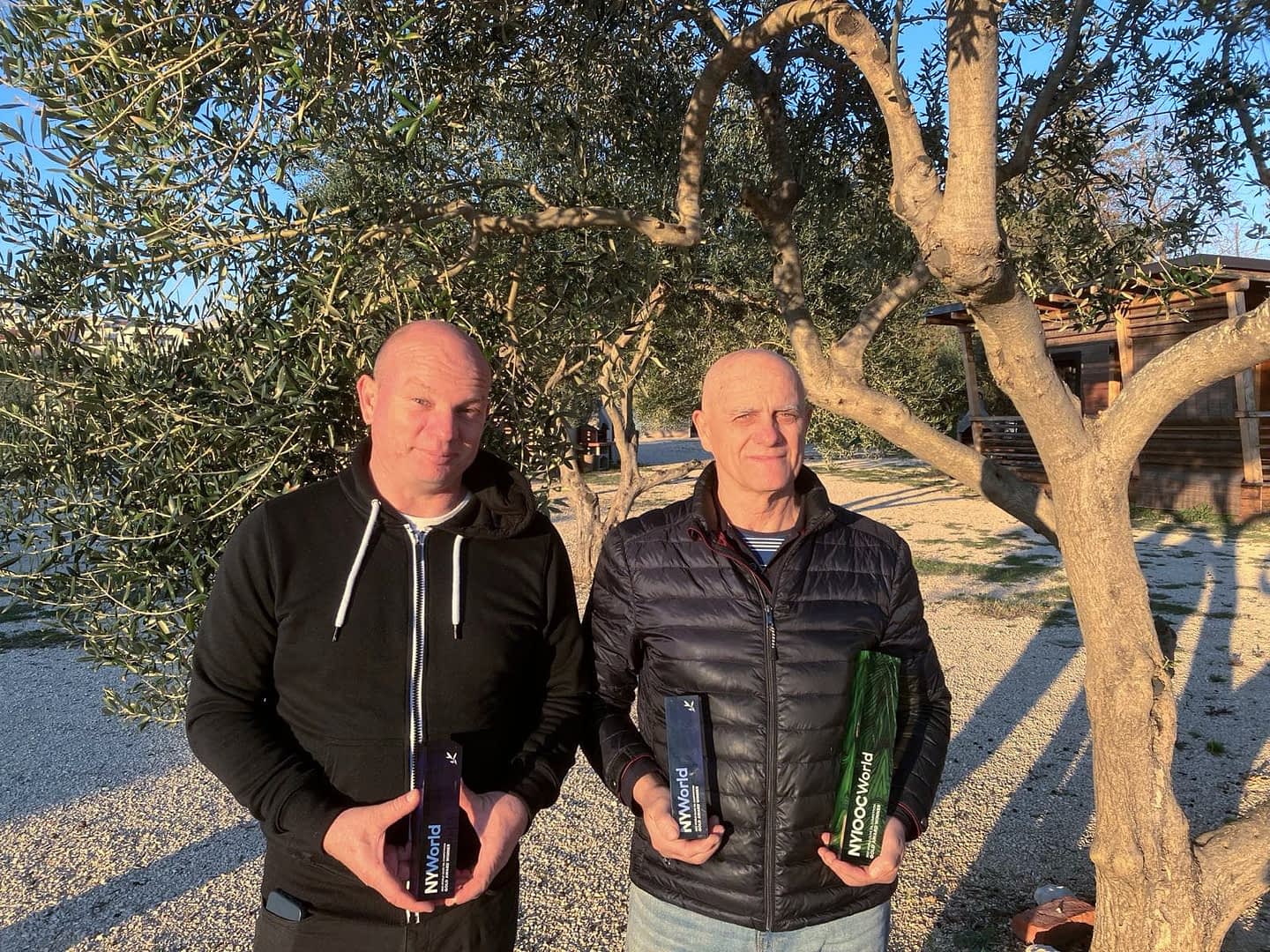 Two men standing outdoors holding bottles of olive oil in front of an olive tree. - Olive Oil Times