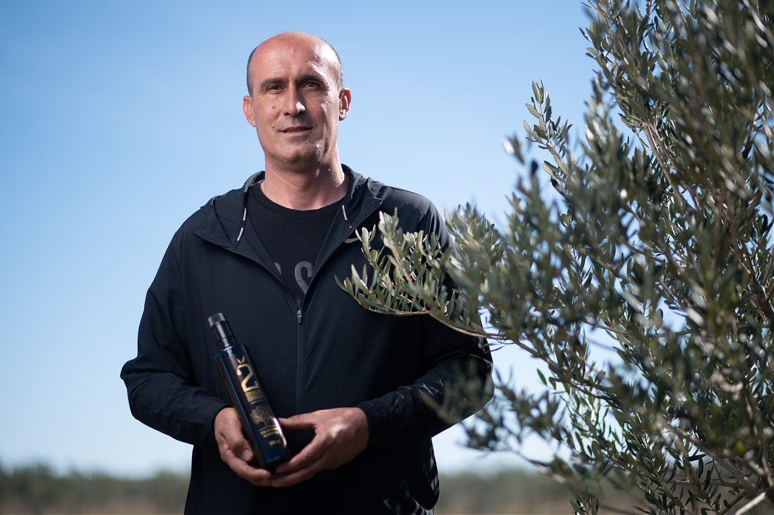 Moški stoji na prostem in drži steklenico olivnega olja poleg oljčnega drevesa. - Olive Oil Times