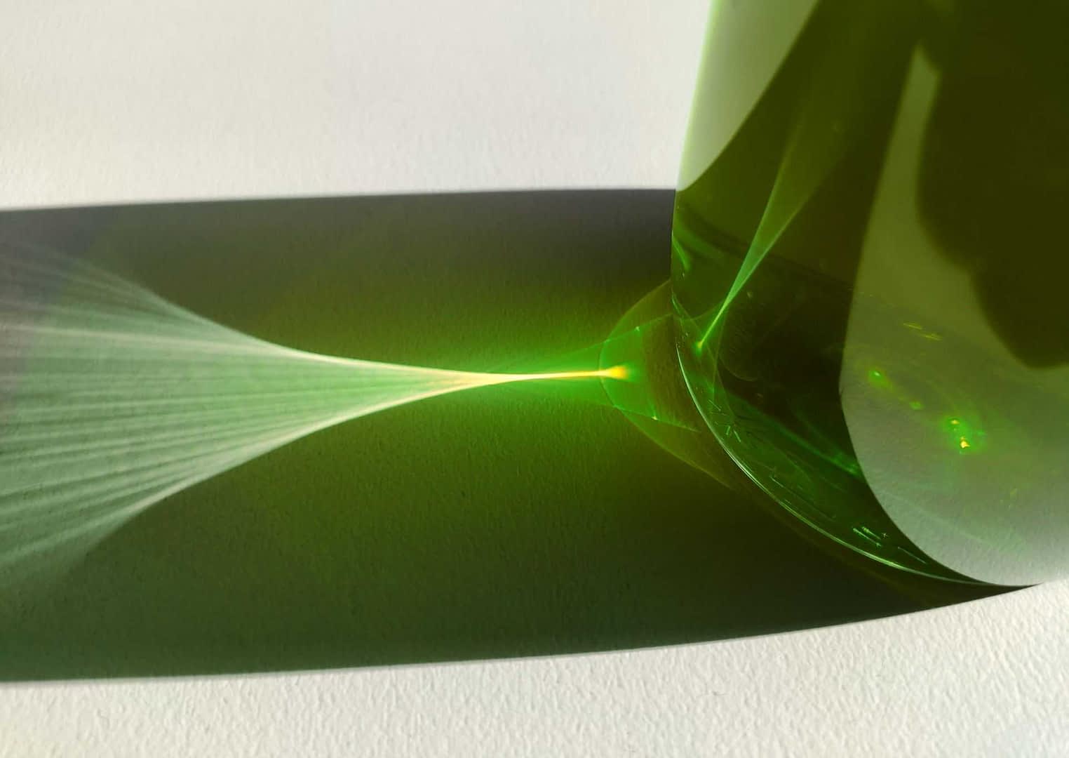 Green glass bottle casting a light reflection on a white surface. - Olive Oil Times