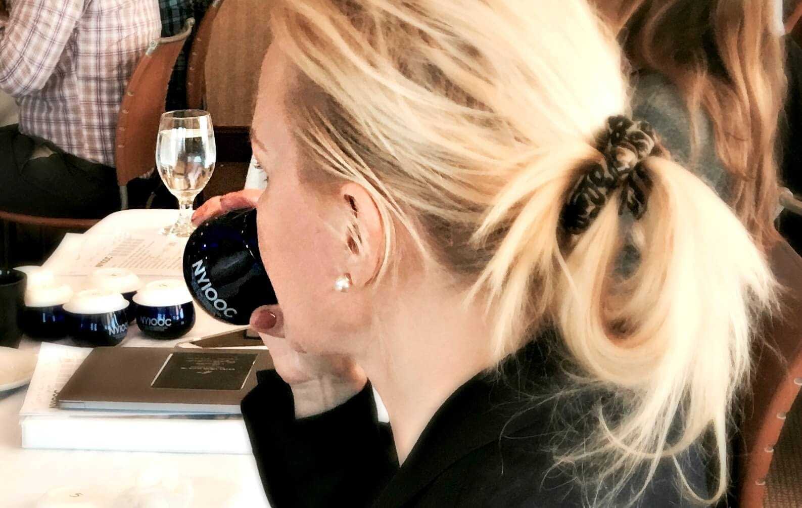 A woman with blonde hair tied in a ponytail holding a blue cup while seated at a table. - Olive Oil Times