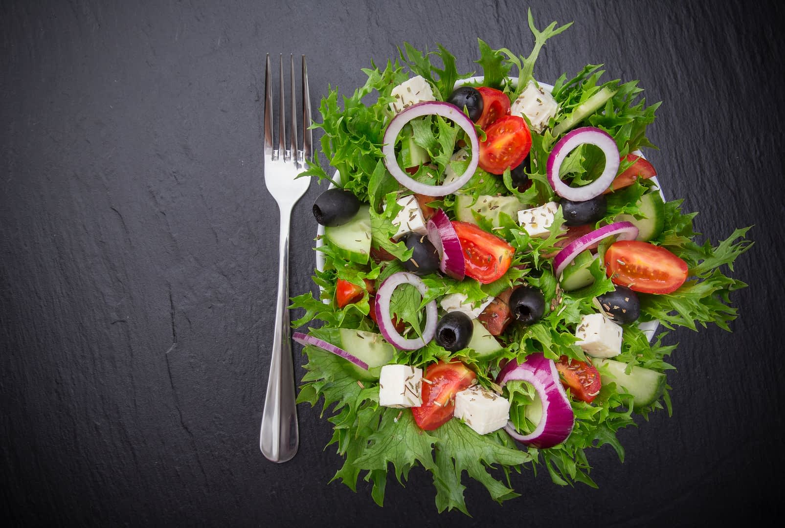 A colorful garden salad featuring mixed greens, tomatoes, cucumbers, olives, and feta cheese on a dark surface. - Olive Oil Times