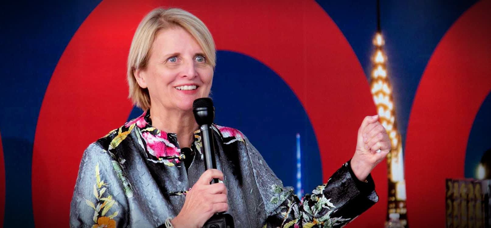 Une femme aux cheveux blonds courts parlant dans un microphone lors d'un événement, portant une veste à motifs floraux. - Olive Oil Times