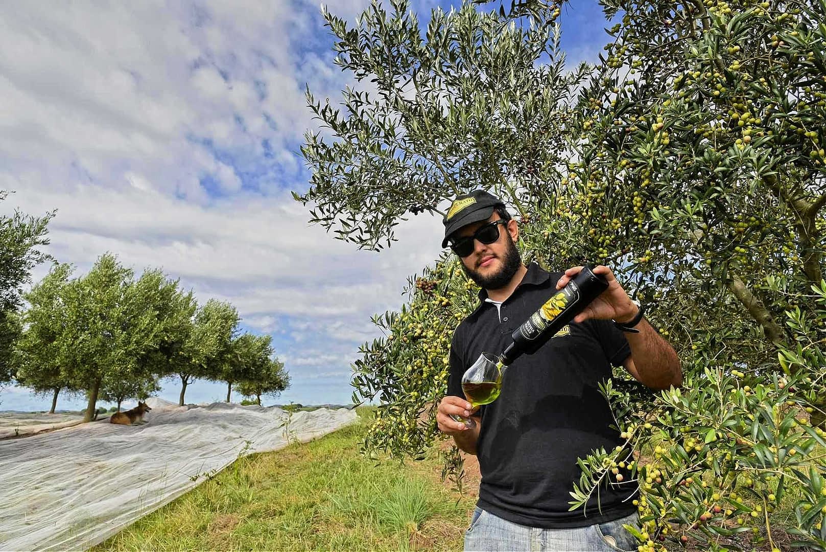 Un hombre con gafas de sol vierte aceite de oliva de una botella en un vaso en un olivar. Olive Oil Times
