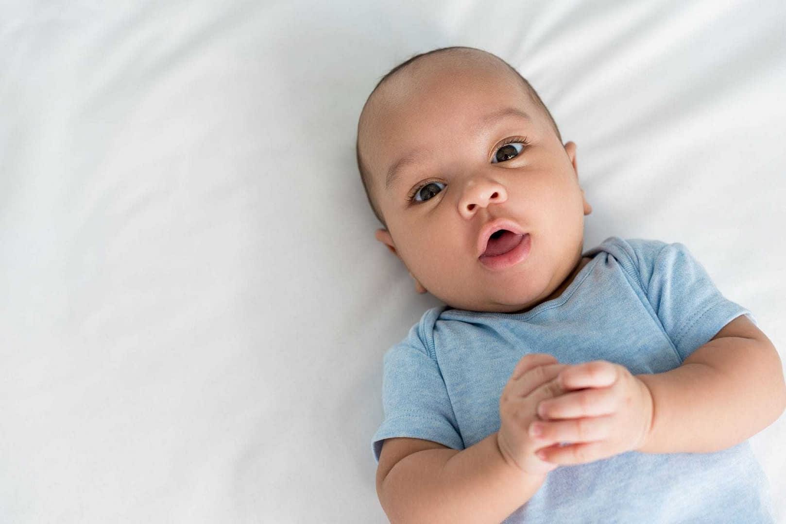 A baby with short hair and a blue shirt lying on a white surface, looking directly at the camera. - Olive Oil Times