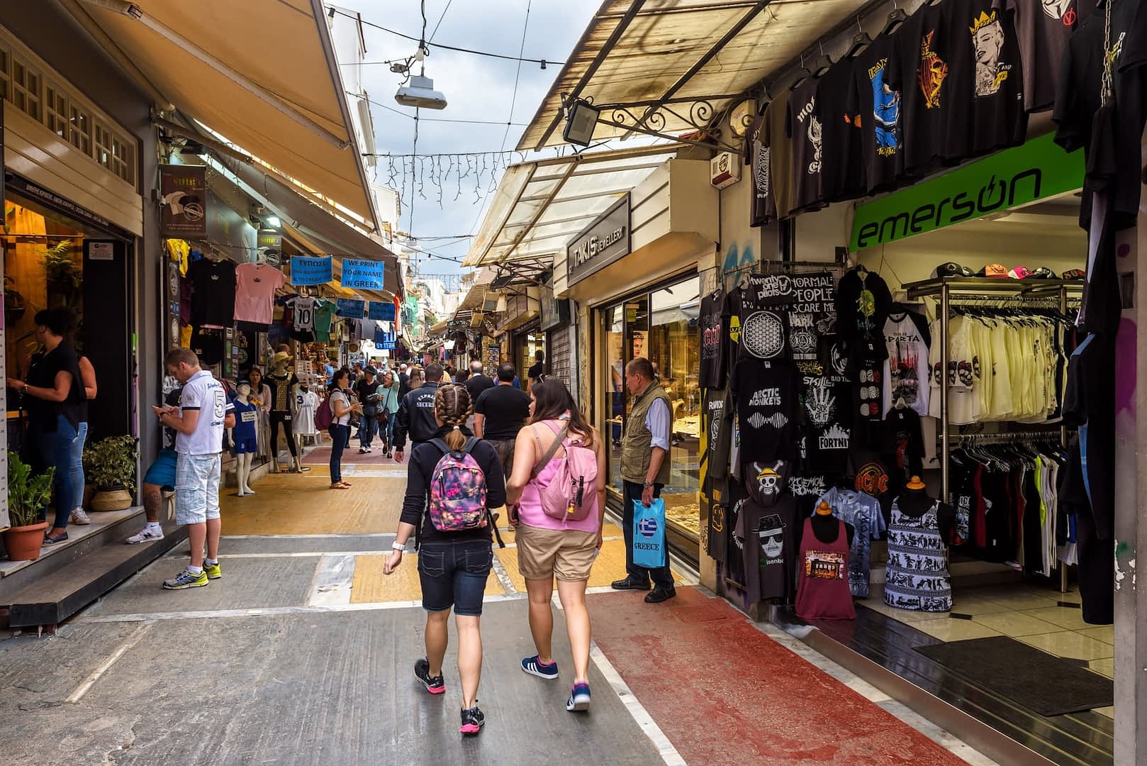 Two people walking through a market street lined with shops and stalls selling various items. - Olive Oil Times