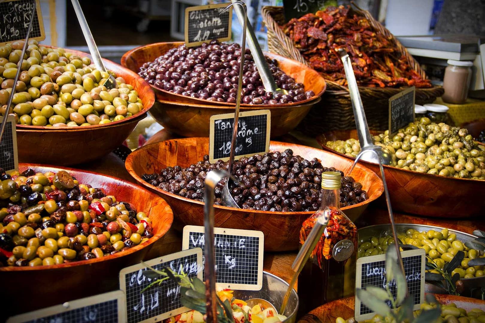 Aceitunas variadas expuestas en cuencos de madera en un mercado, con etiquetas que indican los precios. Olive Oil Times