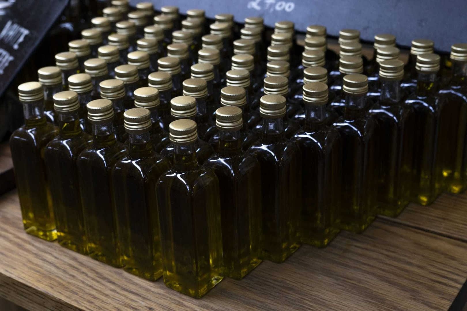 Row of small glass bottles filled with olive oil, each with a gold cap, arranged neatly on a wooden surface. - Olive Oil Times
