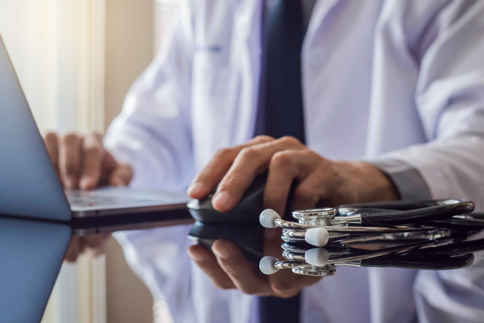 Un professionnel de la santé en blouse blanche utilisant un ordinateur portable avec un stéthoscope sur la table. - Olive Oil Times