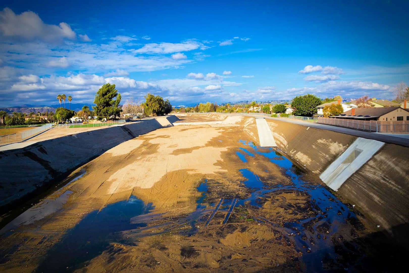 A dry riverbed with patches of water and sparse vegetation under a blue sky with clouds. - Olive Oil Times