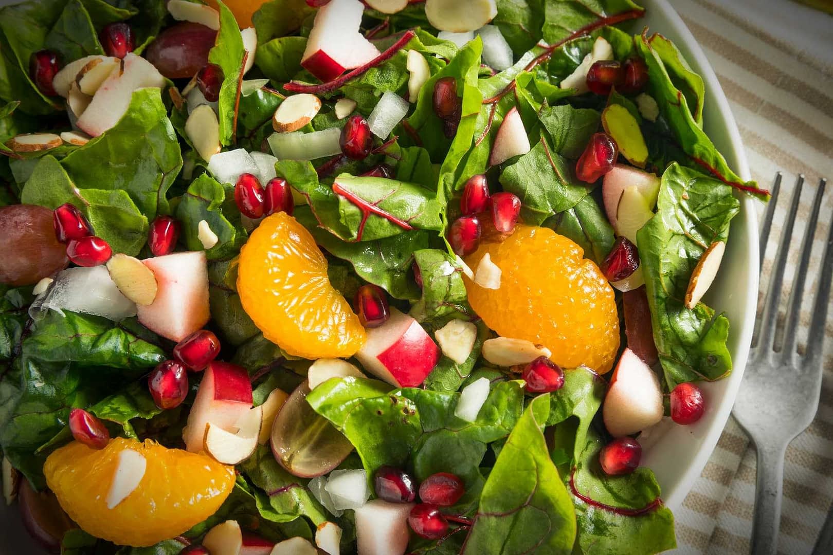 A bowl of mixed green salad featuring spinach, apple pieces, orange segments, pomegranate seeds, and sliced almonds. - Olive Oil Times