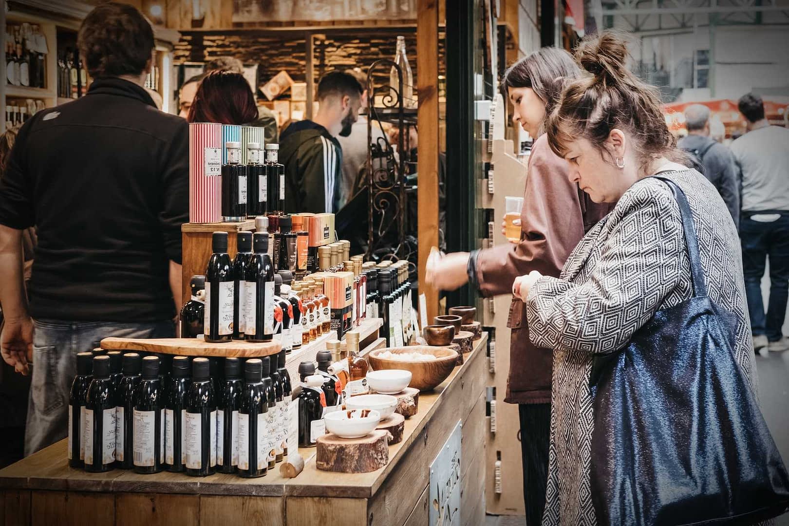 Various bottles of olive oil displayed on a wooden table at a market with people in the background. - Olive Oil Times