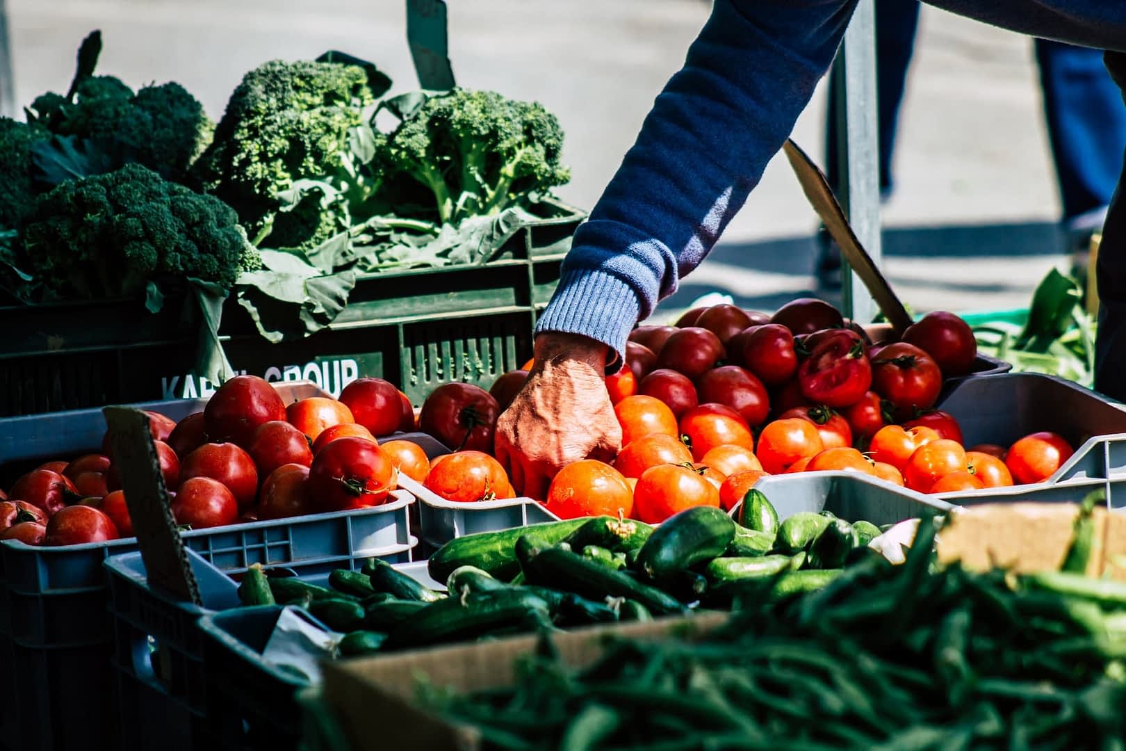 A hand reaching for ripe red tomatoes at a market stall filled with fresh vegetables. - Olive Oil Times