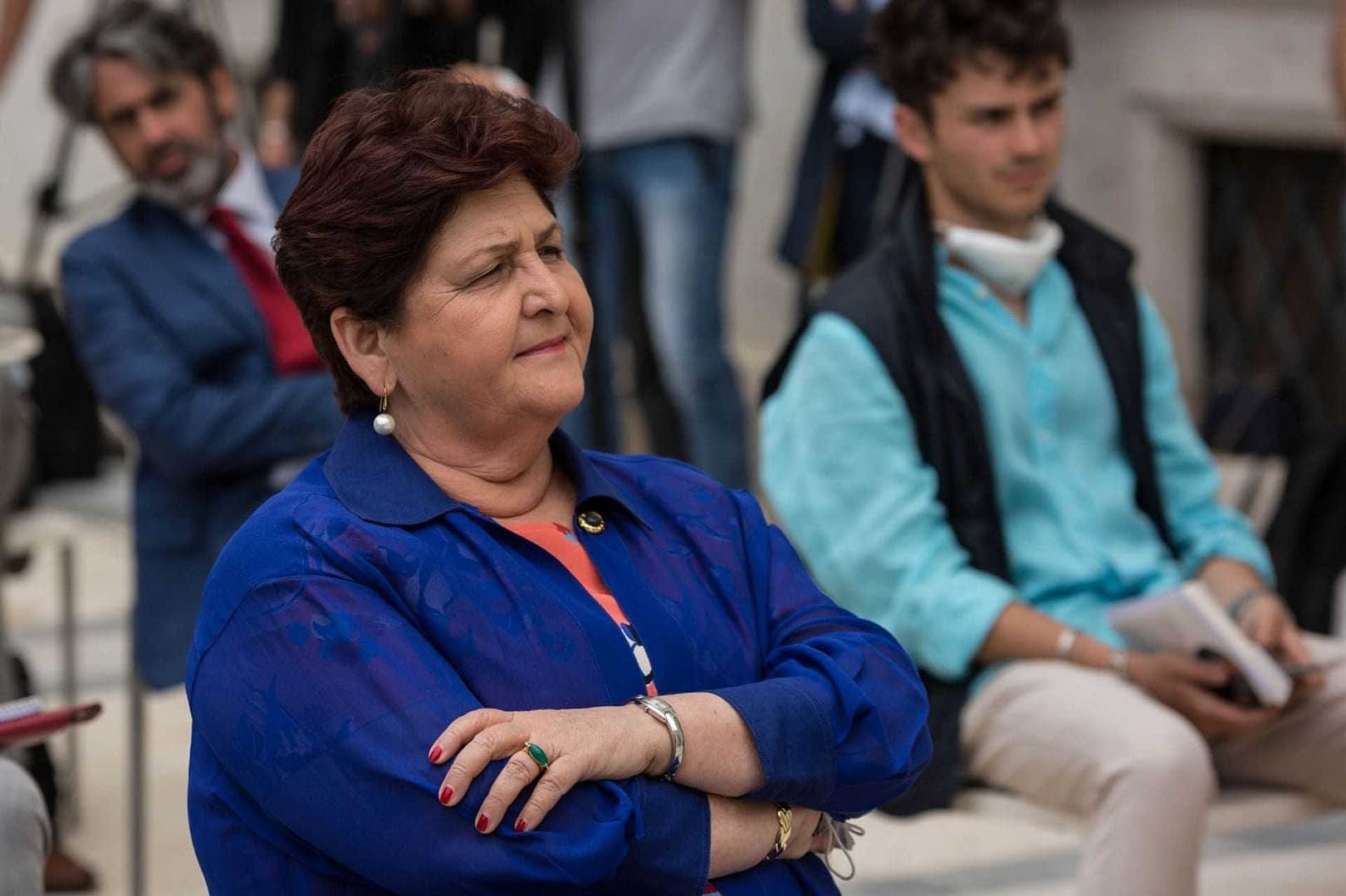Woman with crossed arms wearing a blue jacket and earrings, seated in a group setting with other individuals in the background. - Olive Oil Times