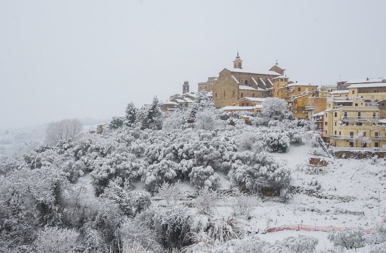 A snow-covered village with buildings and trees blanketed in white snow on a hillside. - Olive Oil Times