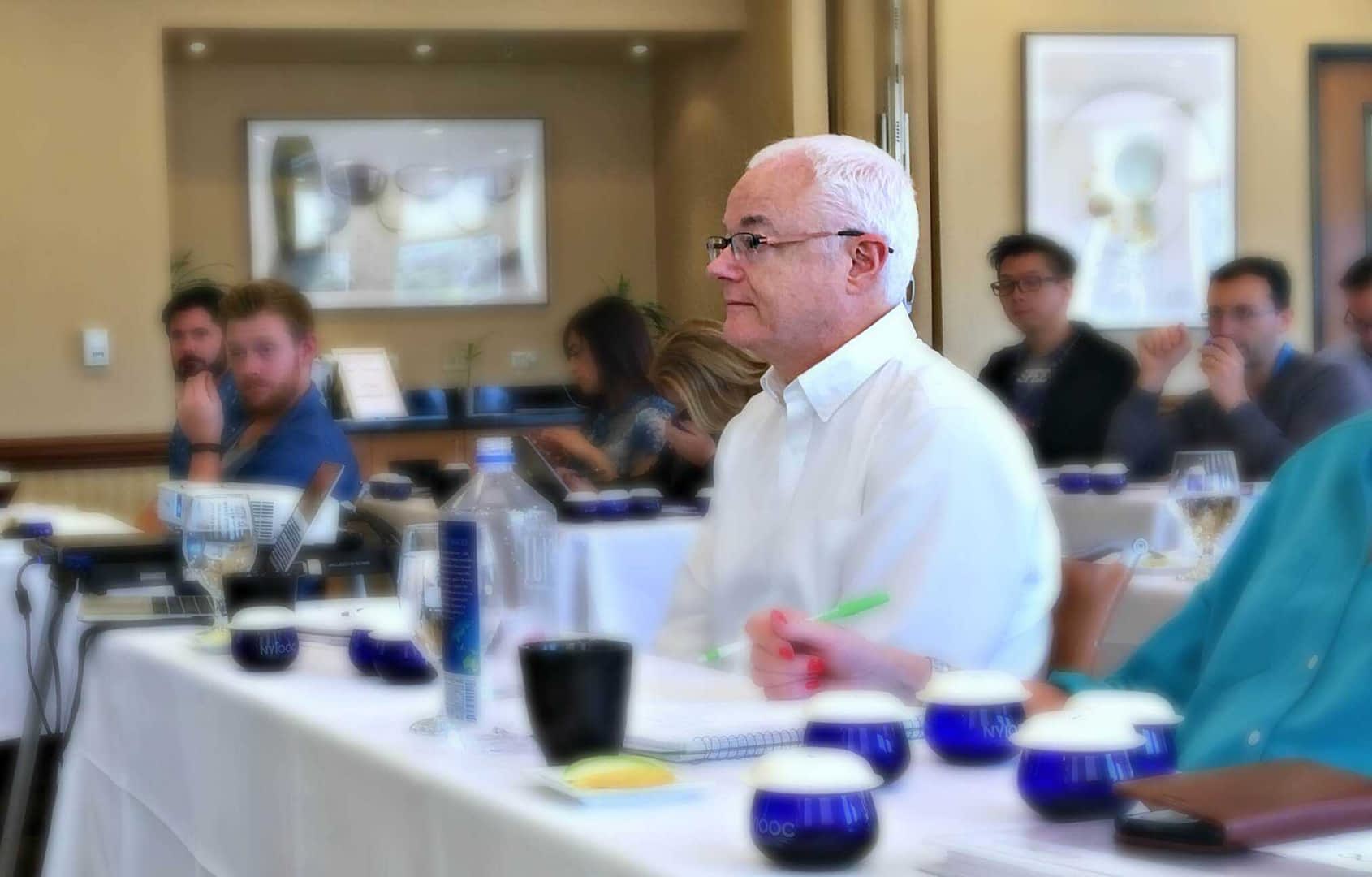 Man with white hair seated at a conference table with participants in the background during a meeting. - Olive Oil Times