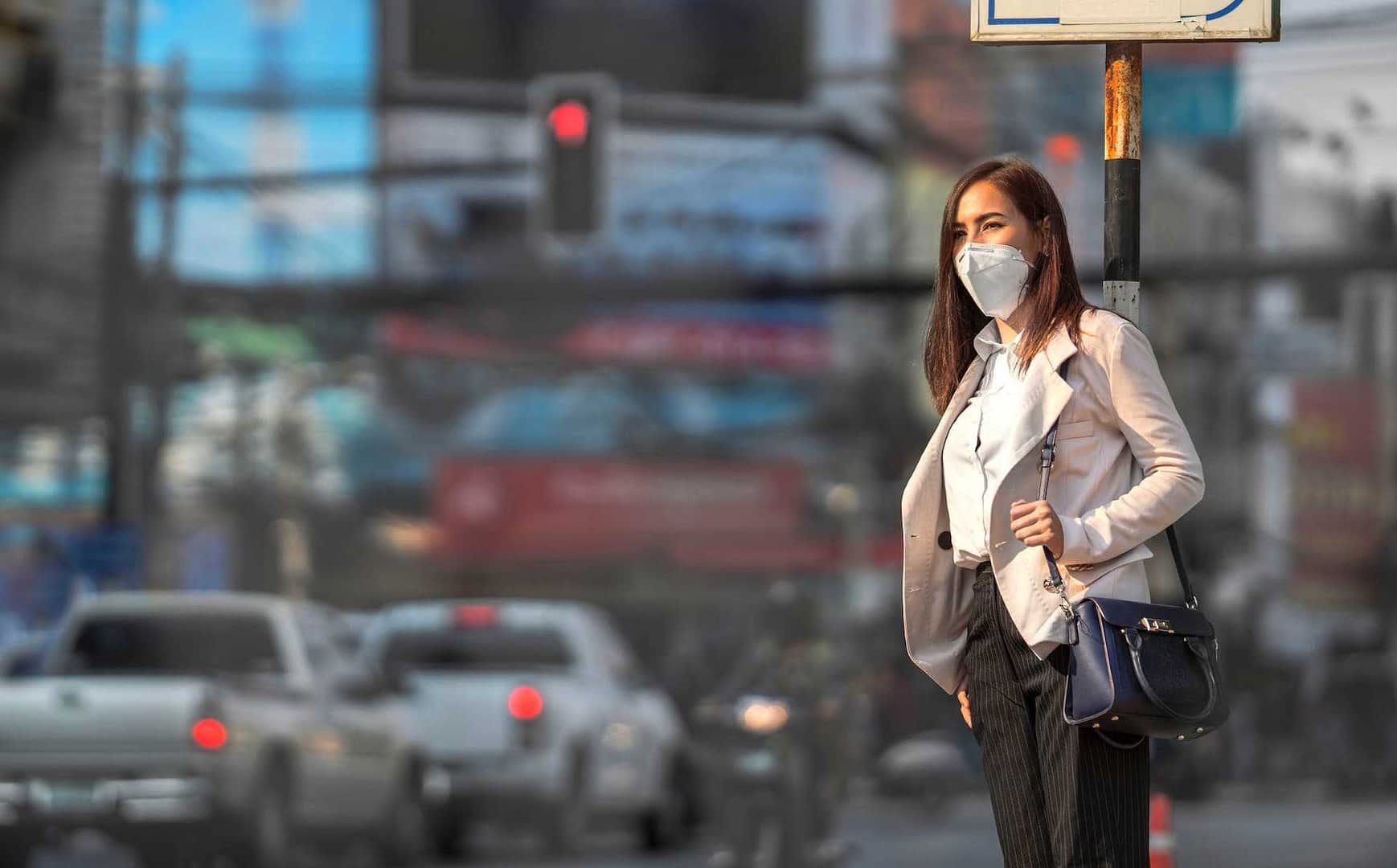 Woman wearing a face mask and business attire standing at a traffic signal in an urban setting. - Olive Oil Times