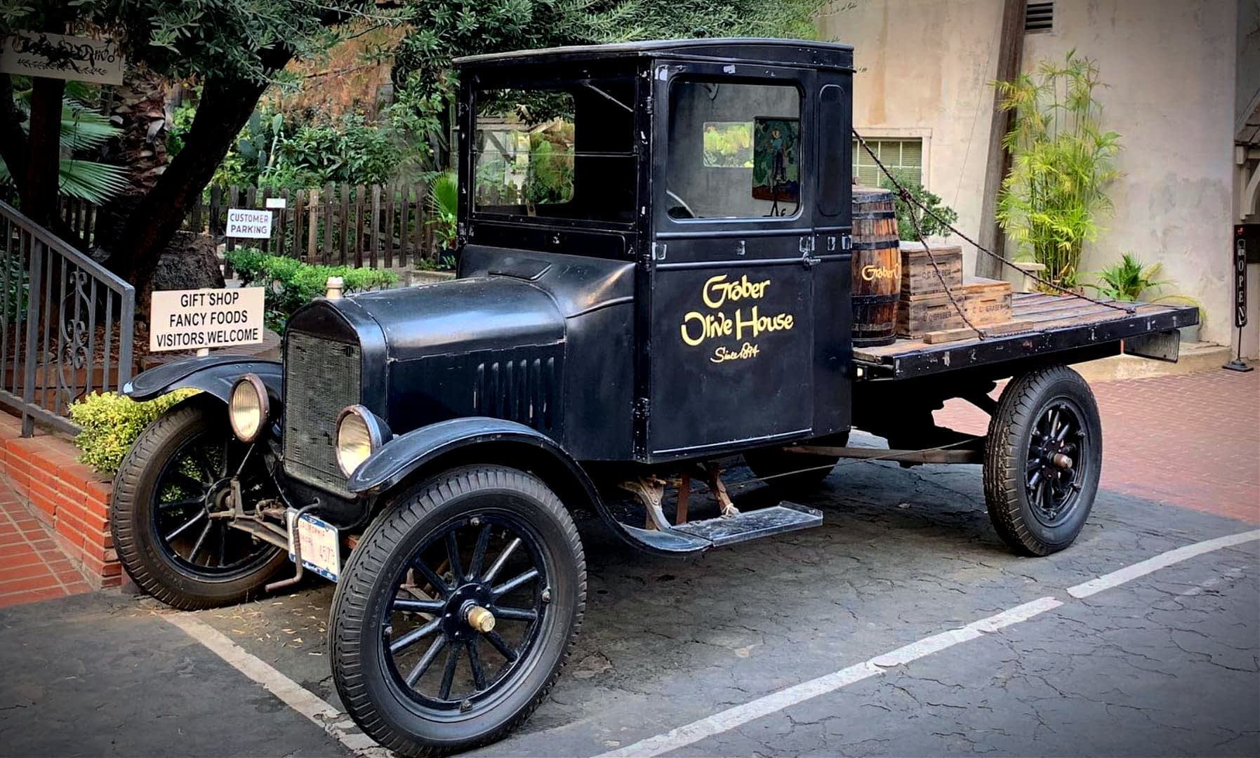 A vintage black pickup truck with wooden barrels in the back, parked on a paved surface. - Olive Oil Times