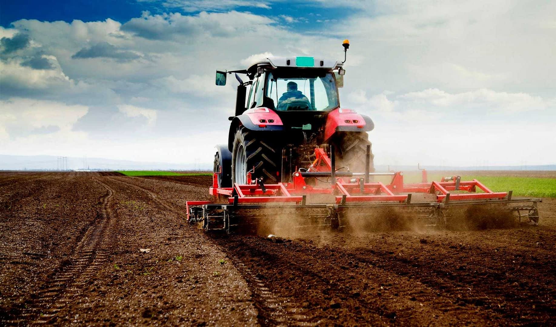 Trattore rosso al lavoro in un campo, arando il terreno mentre si solleva polvere. - Olive Oil Times