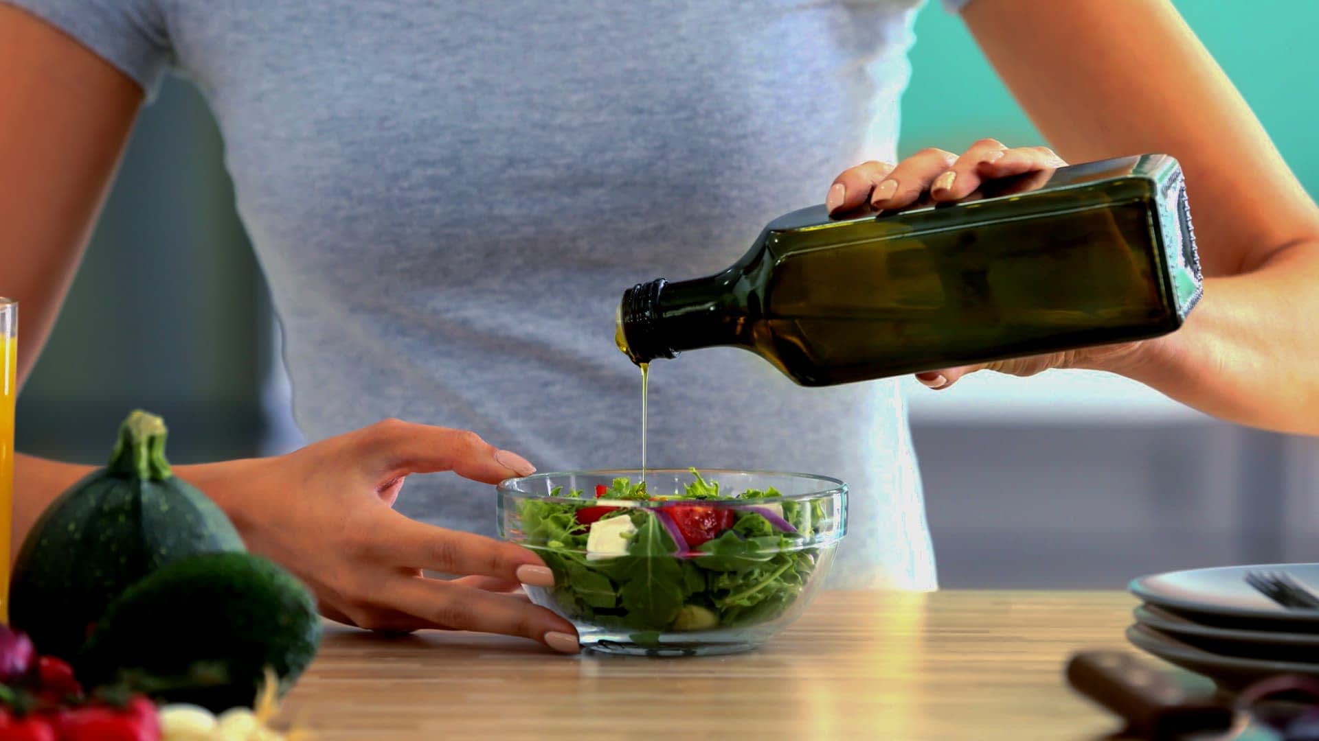 Persona che versa l'olio d'oliva da una bottiglia di vetro scuro su un'insalata fresca in una ciotola trasparente. Olive Oil Times