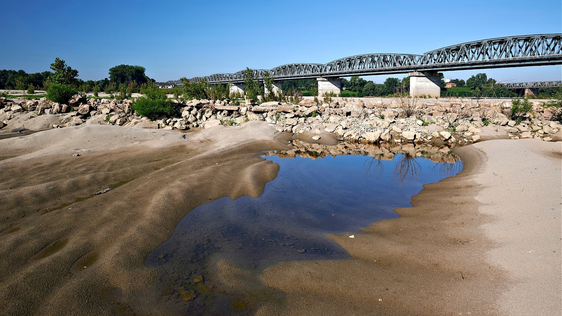 Un puente metálico cruza una ribera de río con terreno arenoso y un pequeño charco de agua en primer plano. Olive Oil Times