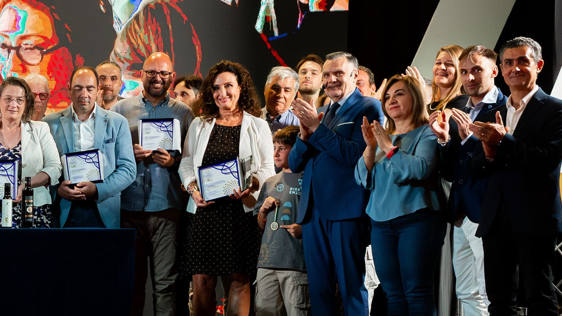 Grupo de personas en una ceremonia de premiación, algunos sosteniendo certificados y aplaudiendo. Olive Oil Times