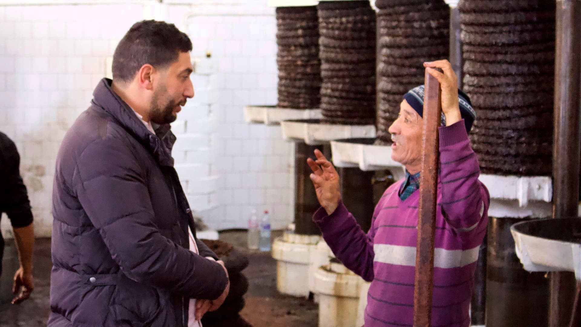 Twee mannen praten in een olieproductiefaciliteit, met apparatuur op de achtergrond. Olive Oil Times