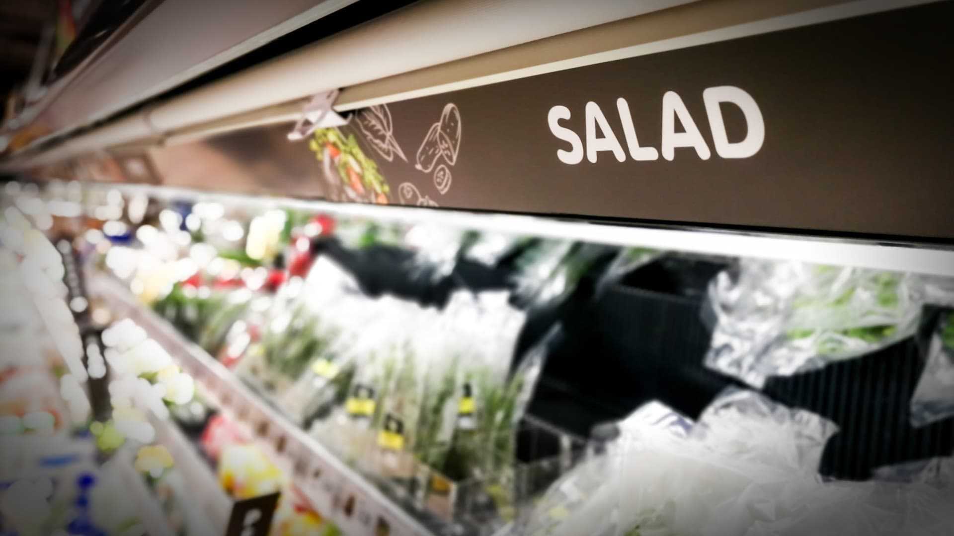 Cartel con la etiqueta 'ENSALADA' en el pasillo de un supermercado con varios ingredientes de ensalada visibles. Olive Oil Times