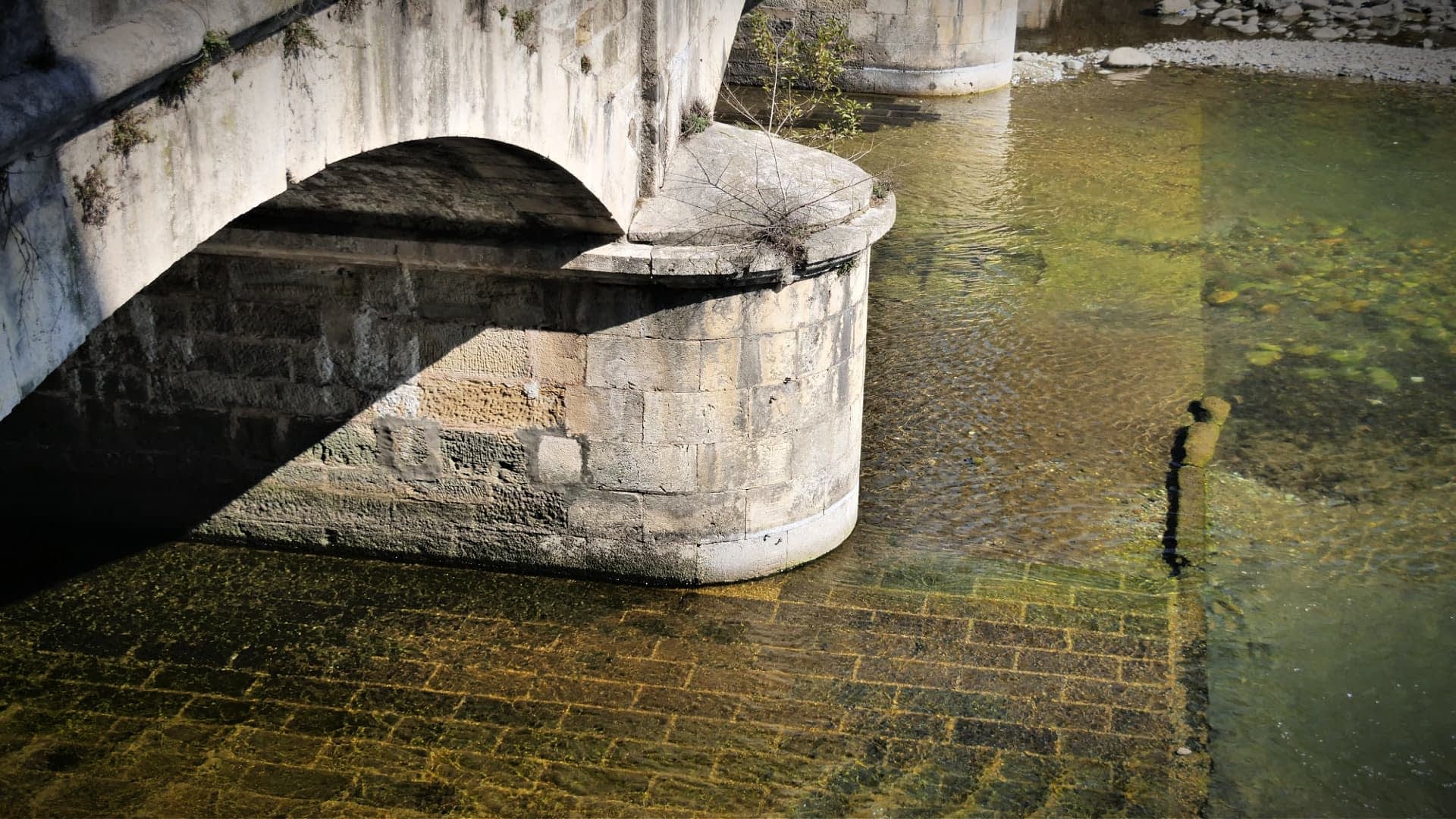 Vista de cerca de un puente de piedra arqueado sobre un cuerpo de agua tranquila con texturas y reflejos visibles. - Olive Oil Times