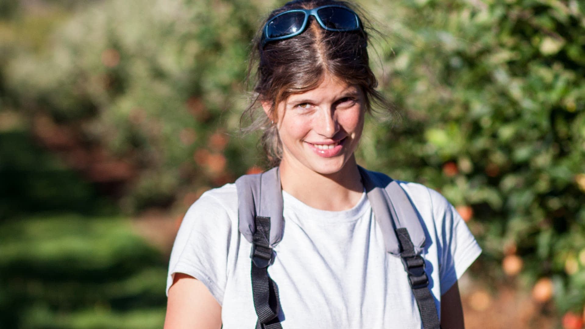 Una donna con occhiali da sole e zaino sorride mentre è in piedi in un frutteto con alberi sullo sfondo. - Olive Oil Times
