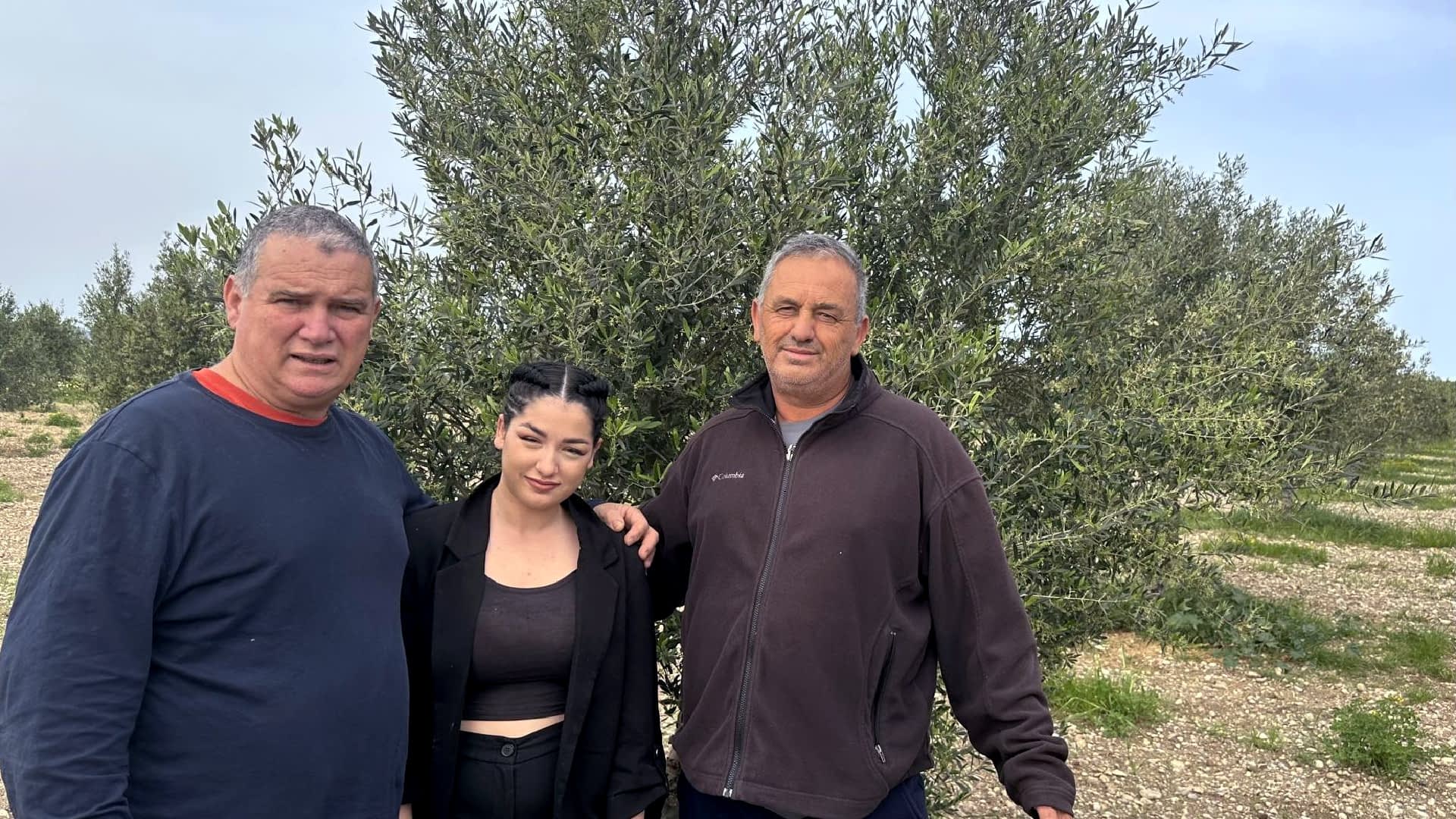 Drie personen staan ​​samen voor een olijfboom in de buitenlucht. Olive Oil Times