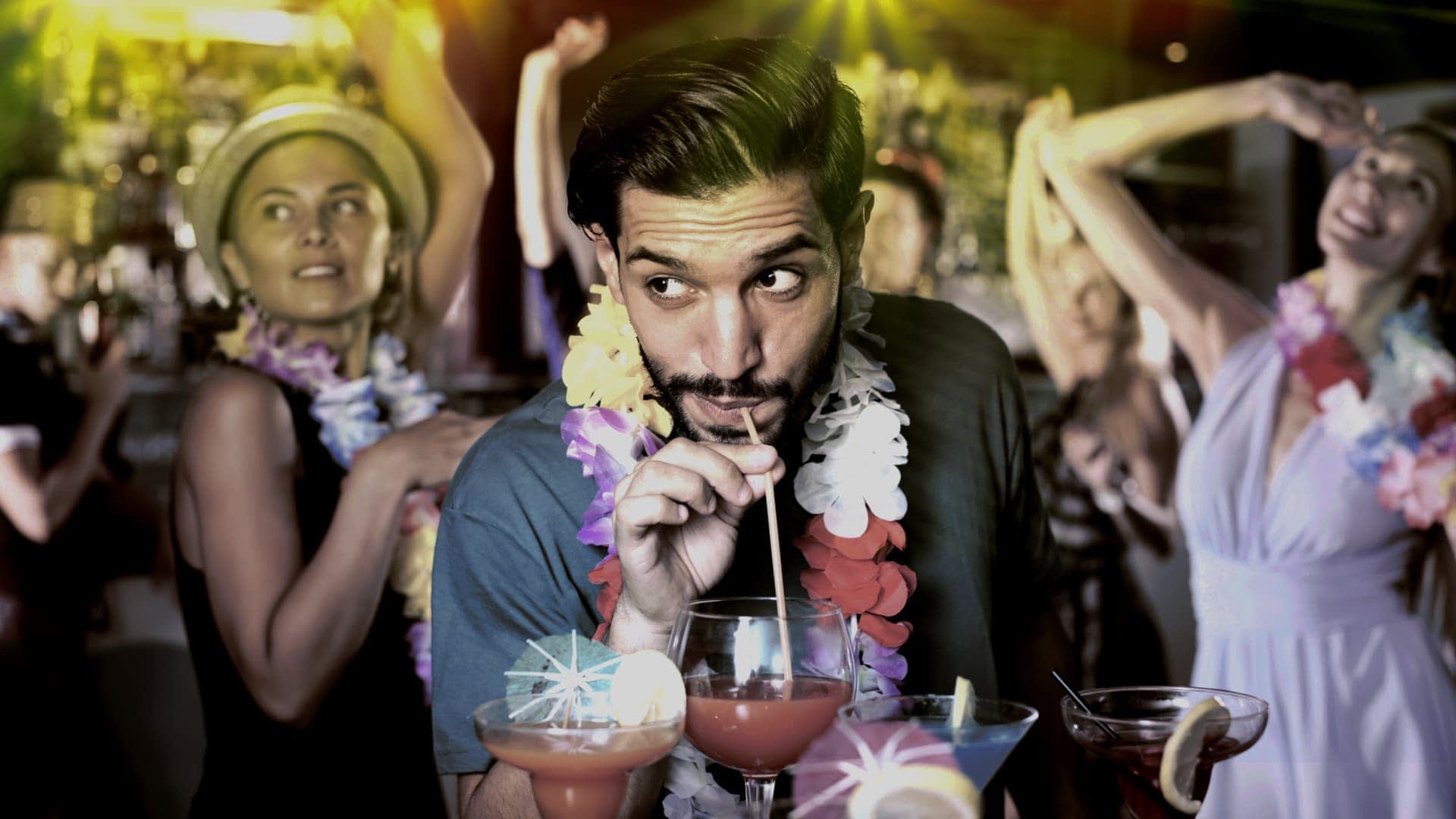 Man sipping a cocktail through a straw while surrounded by people at a party. - Olive Oil Times