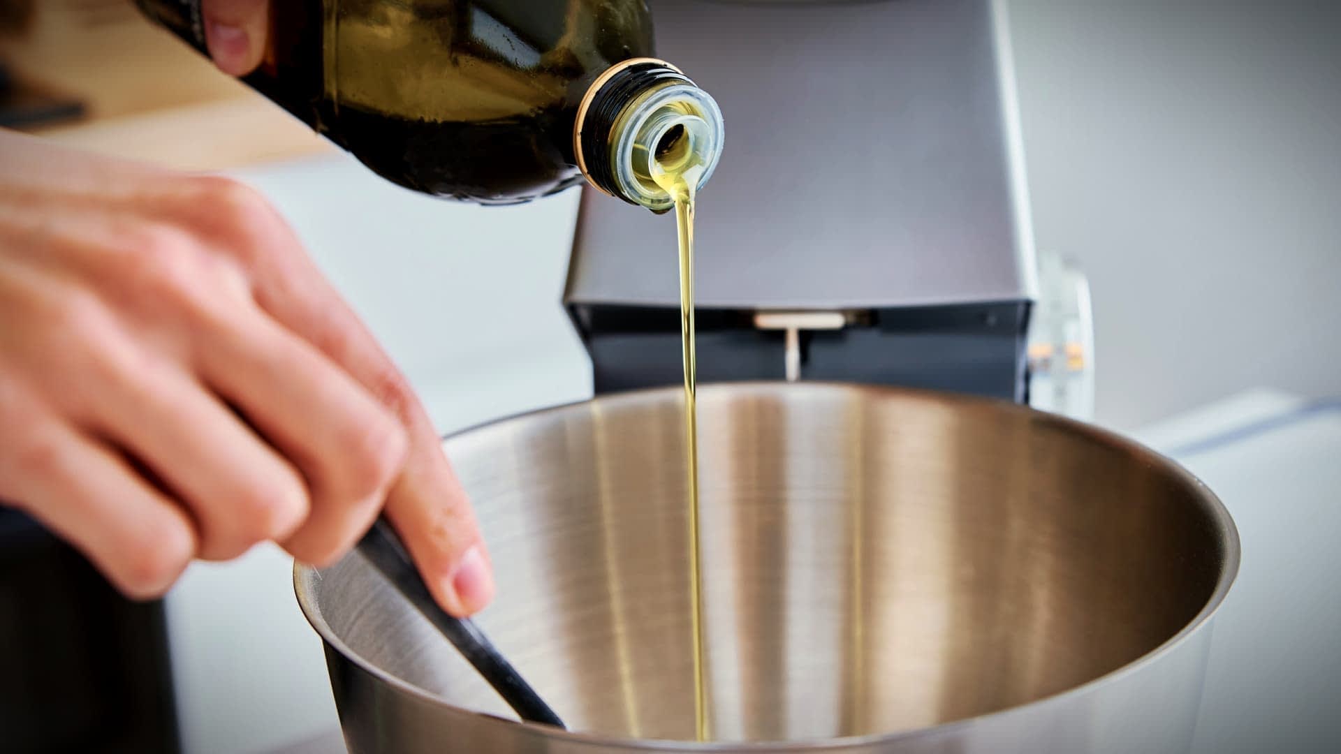 Une personne versant de l'huile d'olive d'une bouteille dans un bol à mélanger en acier inoxydable. - Olive Oil Times