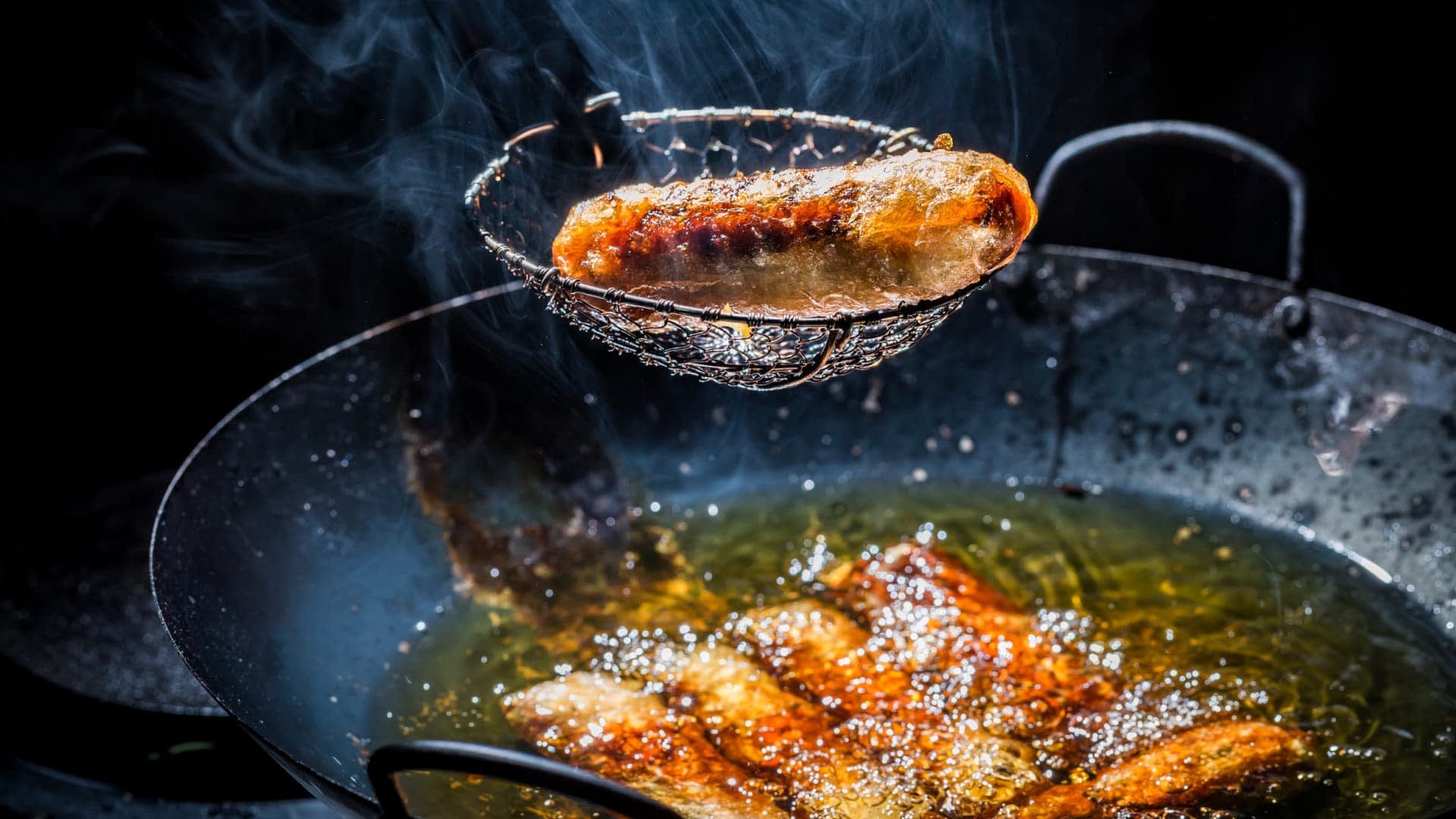 Une passoire en métal soulevant une saucisse frite d'une poêle d'huile chaude avec une surface bouillonnante. - Olive Oil Times