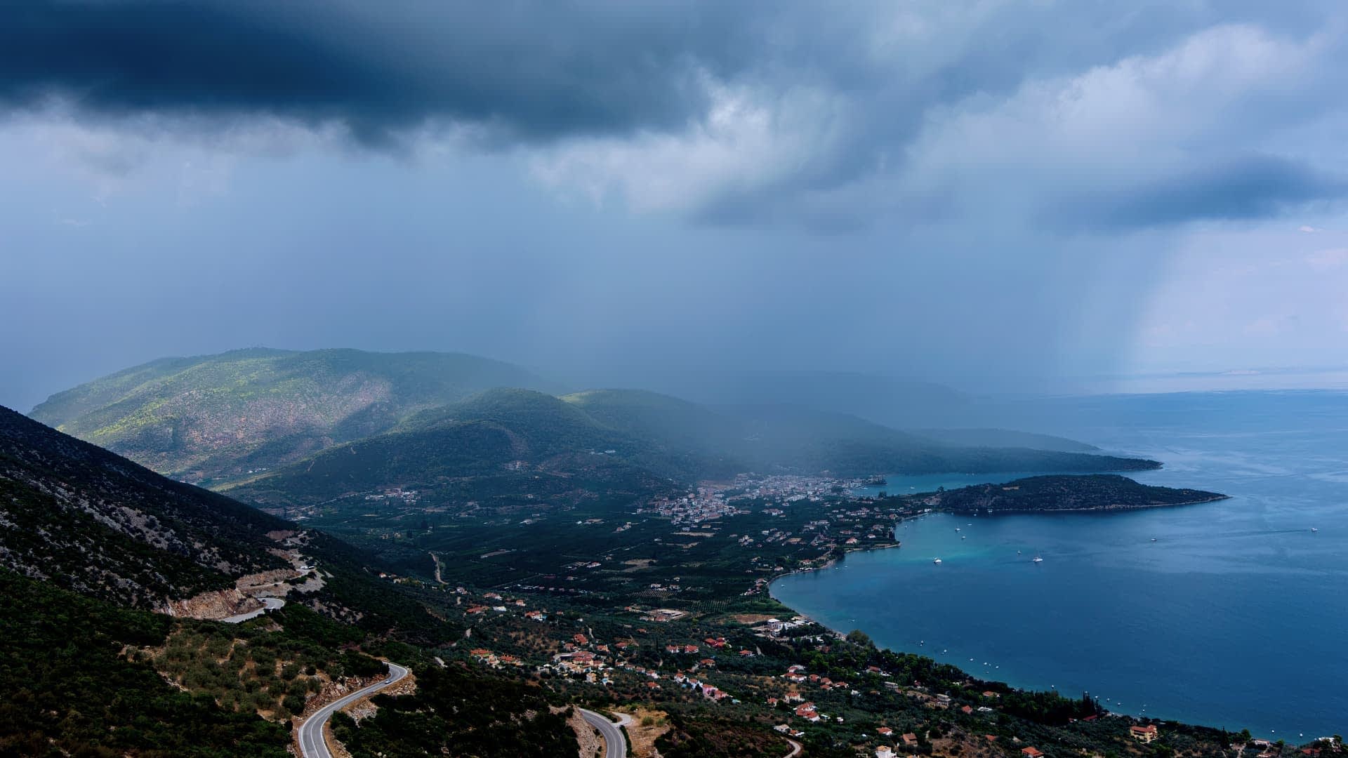 Dağların, bir koyun ve suyun üzerinde bulutlu gökyüzünün yer aldığı bir kıyı manzarası. - Olive Oil Times