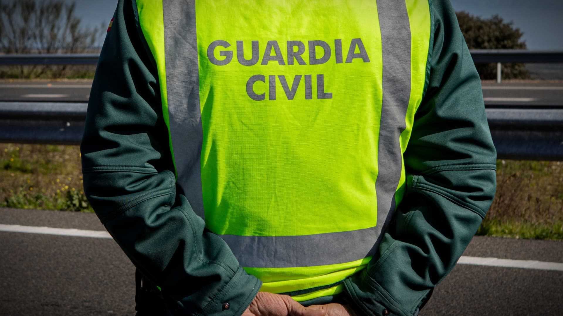 Офицер Гражданской гвардии в светоотражающем жилете стоит на обочине дороги, заложив руки за спину. Olive Oil Times