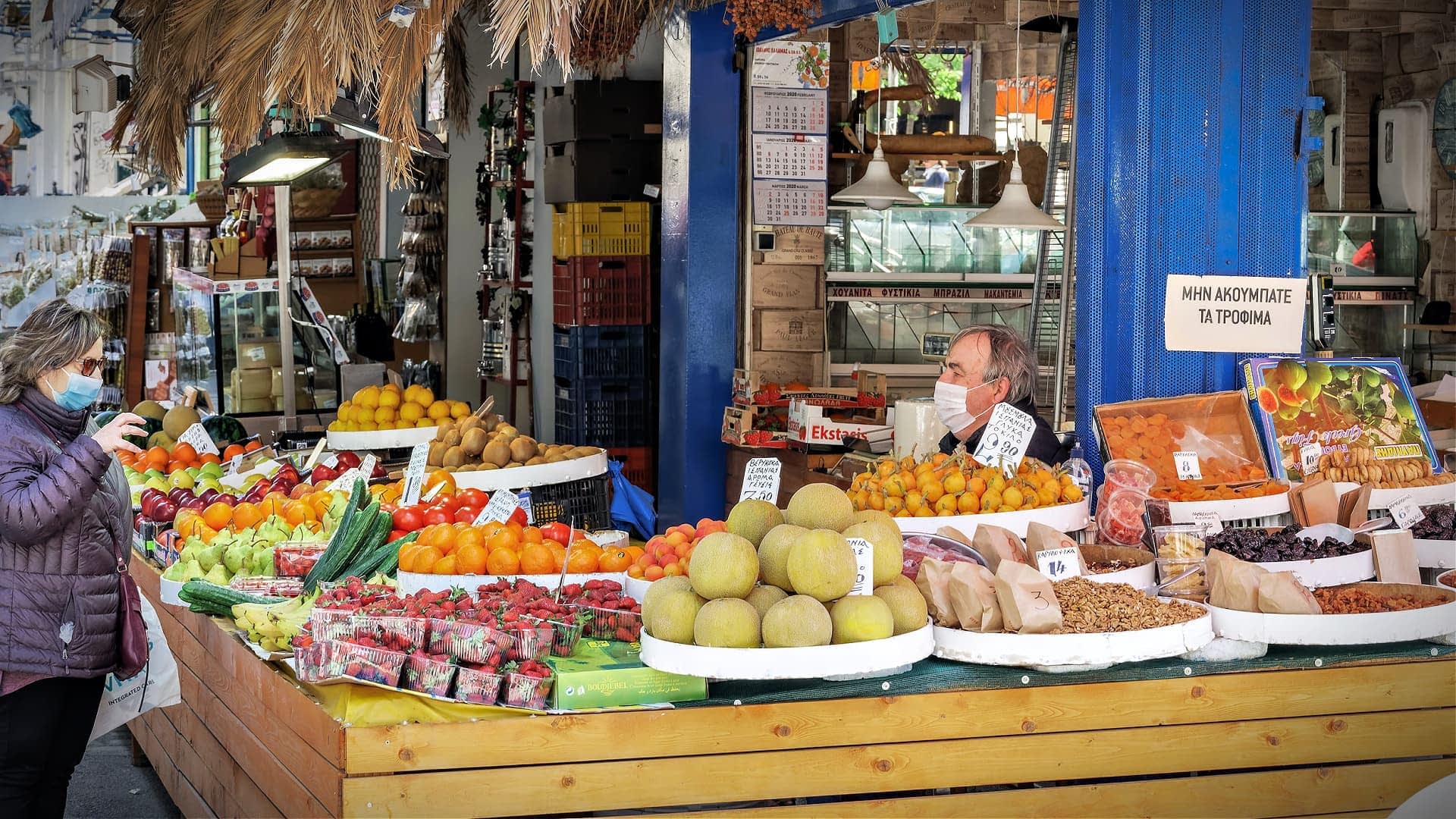 Puesto de mercado que muestra una variedad de frutas y verduras, con un vendedor y un cliente con máscaras. Olive Oil Times