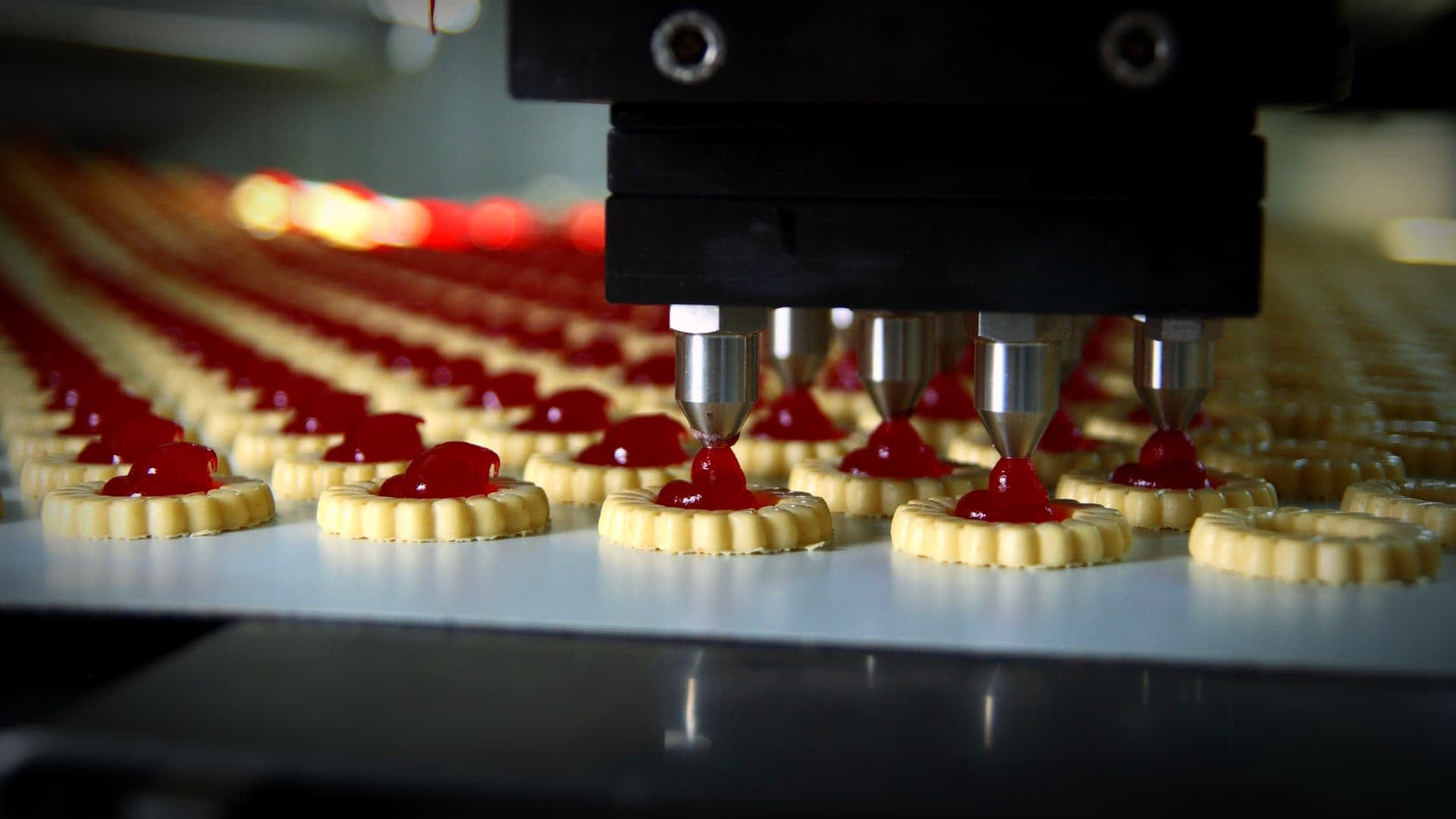 Macchina automatica che distribuisce il ripieno rosso sulle basi dei biscotti in una linea di produzione. Olive Oil Times